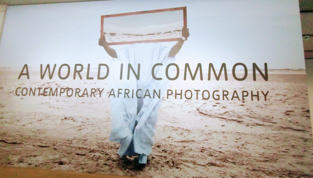 Person holding frame in a dessert or sand. Text of exhibition name, A world in common, contemporary African photography'.