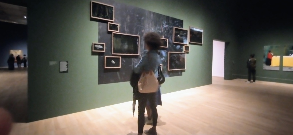 Dark green wall with little frames across a blackish wall. Woman with denim top standing with back to us. 